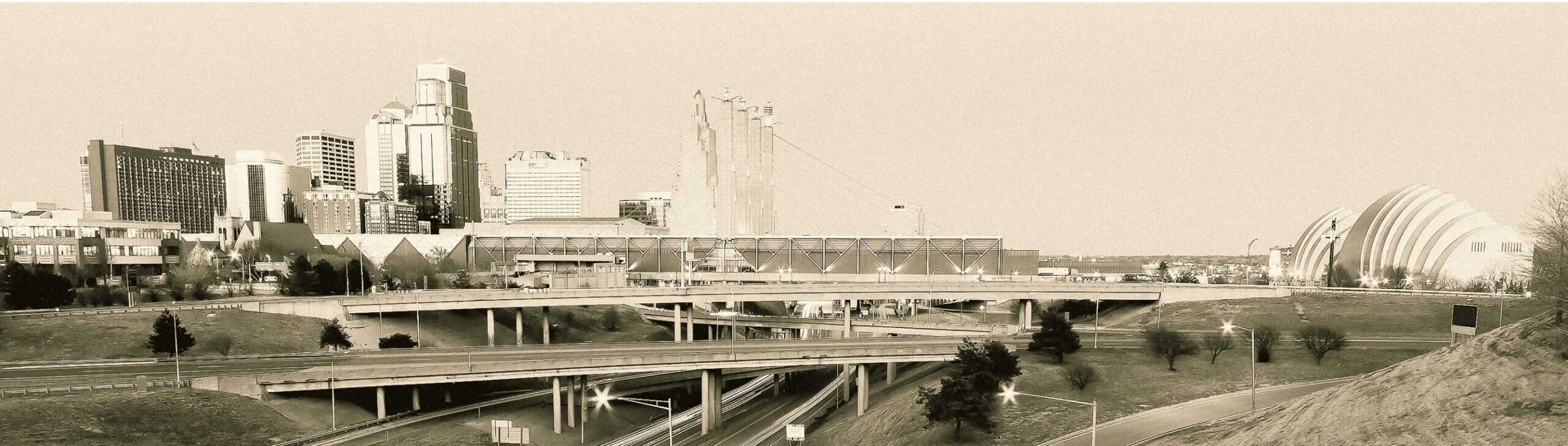Sepia-toned image of a cityscape featuring highways in the foreground and a skyline with various buildings. Prominent structures include a domed building on the right and a tower in the center. Roads intersect below with minimal traffic.