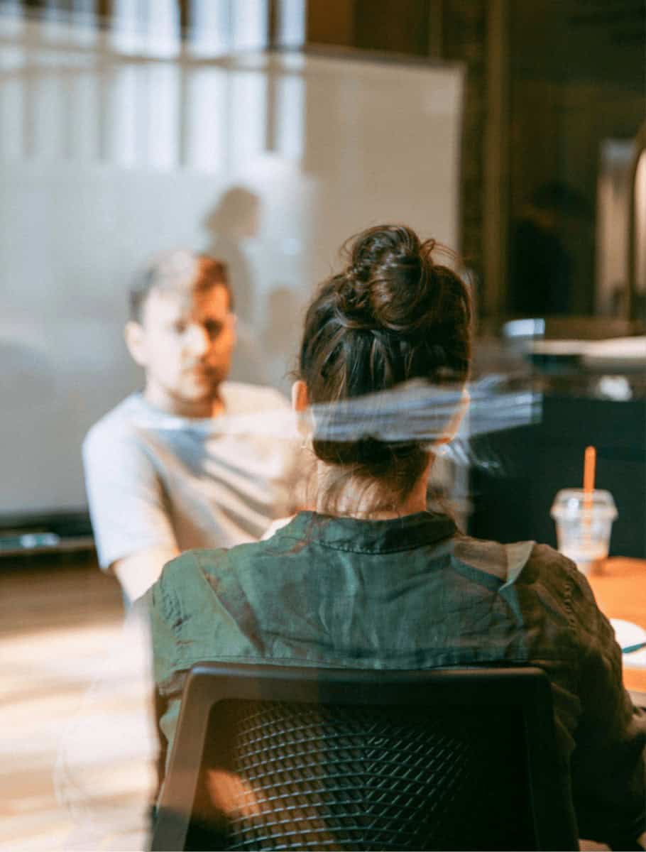 Two people sitting at a table in an office setting, viewed through a glass window. The person closest has their back to the camera, and the other is facing forward. A whiteboard and a plastic cup with a straw are visible in the background.