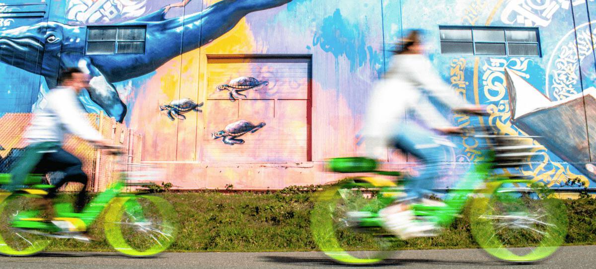Two people ride bright green bikes past a colorful mural featuring sea turtles and oceanic designs on a building. The movement creates a blur effect, contrasting with the vivid artwork.