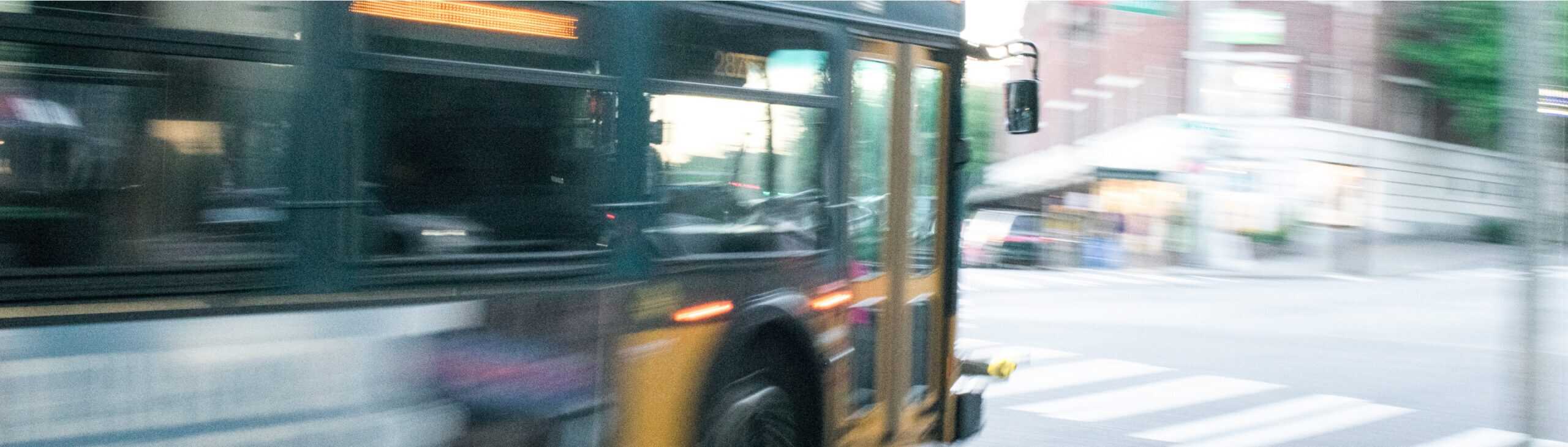 A blurred image of a moving city bus, likely in an urban area. The bus is turning a corner, and there are buildings and a crosswalk in the background. The motion creates a sense of speed and movement.
