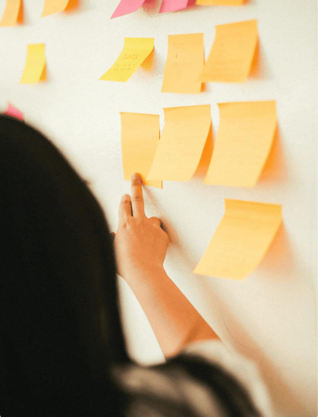 A person with long hair is placing a yellow sticky note on a white wall, surrounded by other yellow and pink sticky notes. Only the persons hand and part of their head are visible. The scene suggests a brainstorming or planning session.