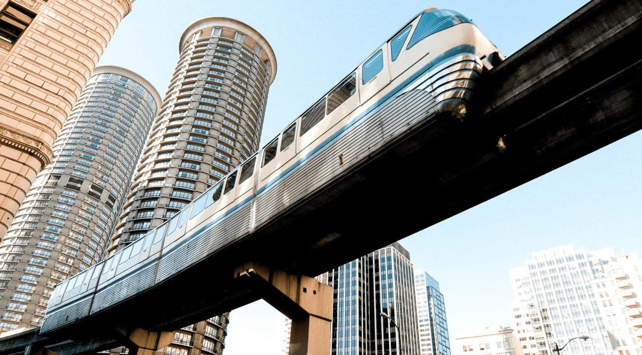 A futuristic monorail train travels on an elevated track in an urban setting, surrounded by tall, cylindrical skyscrapers. The sky is clear, enhancing the modern architectural feel of the scene.