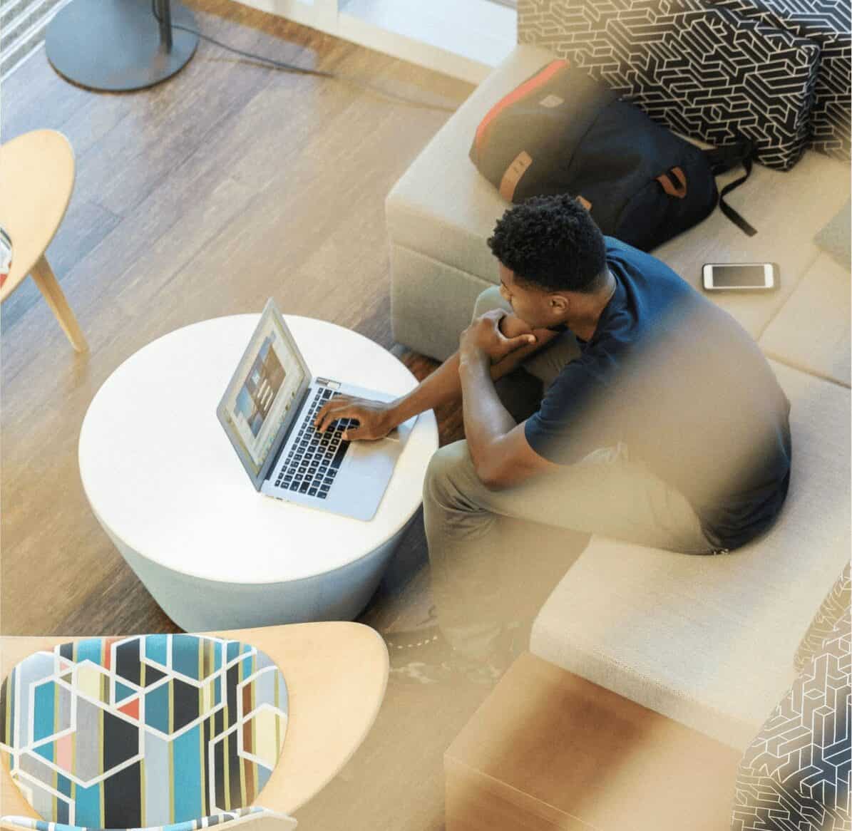 Person sitting on a couch in a modern workspace, using a laptop on a round table. A backpack and smartphone are nearby. Geometric-patterned pillows decorate the space, which has a wooden floor and minimalistic furniture.