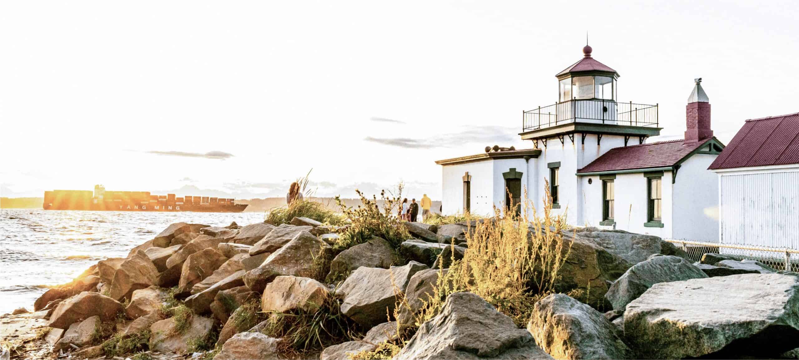 A lighthouse with a red roof stands on a rocky shoreline as the sun sets. A cargo ship is visible in the background on the ocean. The scene is serene, with warm light illuminating the lighthouse and rocks.